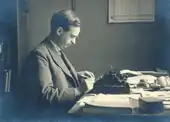Middle-aged Hendrik Coenraad Blöte using a type writer. Hendrik is wearing a suit, tie and jacket. Many papers, some books and a jar of ink are on the desk.