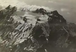 Old photo of a mountain in the clouds with a white glacier at the foot of the peaks.